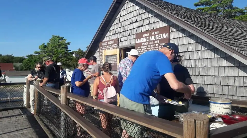 People gather at a food counter beside a shingled museum building.