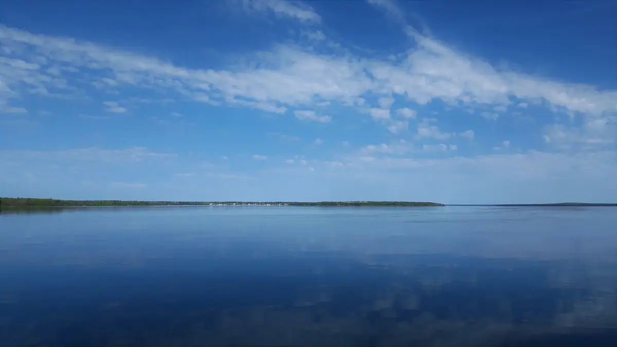 Calm blue water reflects a broad sky above a low, wooded shoreline with a few distant pale structures.