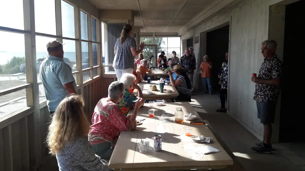 People gather around long tables for a meal or presentation in a screened porch overlooking the shoreline.
