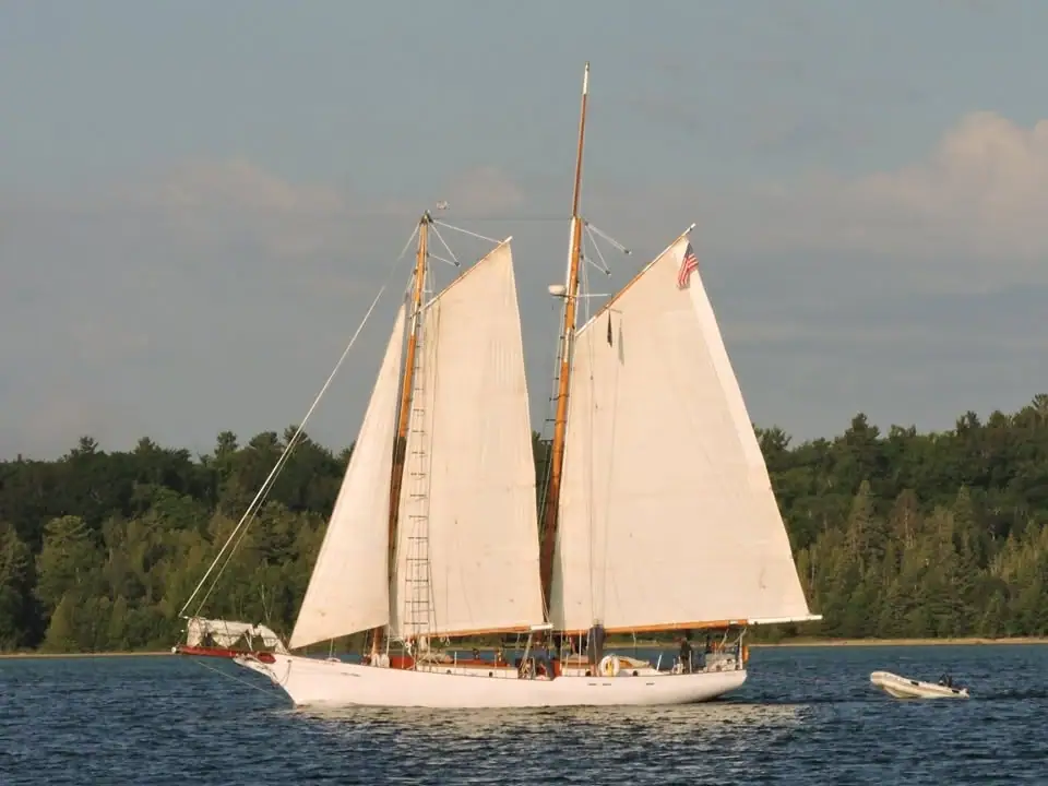 A white two-masted schooner sails past a forested shoreline with a small inflatable boat nearby.