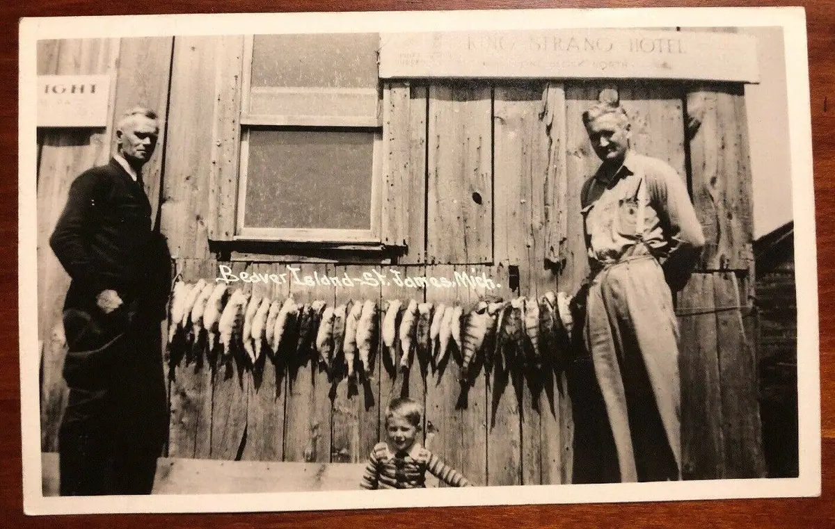 A vintage postcard shows two adults and a child beside a wooden building with animal pelts displayed outside.