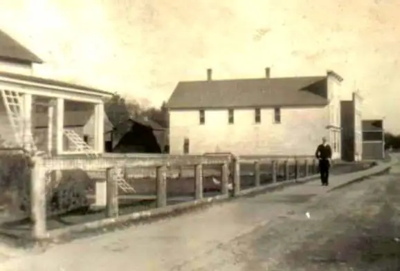 Sepia photograph of a street with a fenced porch, large two-story building, and a person walking.