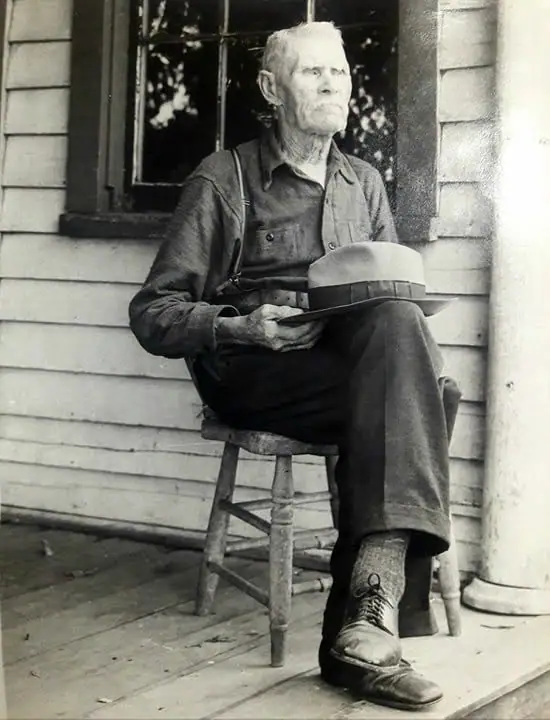 Black-and-white portrait of an older man seated on a porch, holding a light-colored hat across his lap.