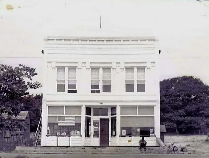 Black-and-white photograph of a two-story storefront with an American flag, trees, and a bicycle outside.