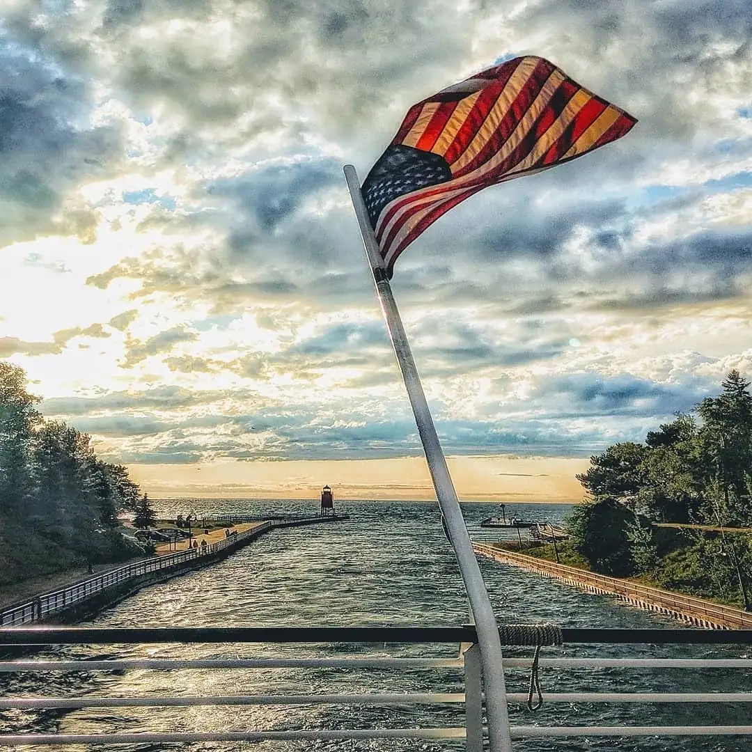An American flag flies above a boat railing as sunlight breaks through clouds over a channel and lighthouse.