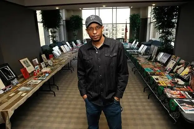 A man stands between two long tables displaying framed photographs, books, magazines, and other items.