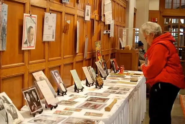 A woman examines photographs and papers arranged on a long display table in a wood-paneled room.