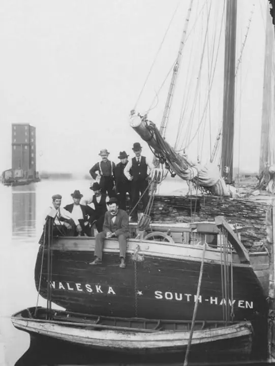 Several men pose aboard a schooner with rigging, stacked lumber, and a small boat alongside.