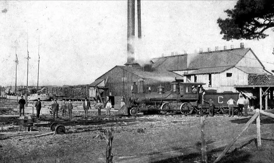 Steam locomotive and workers beside a lumber mill, with stacked timber, rail tracks, and a schooner in the background.
