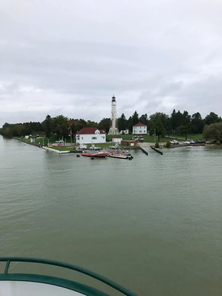 A lighthouse, waterfront buildings, small boats, and a pier seen across calm water.