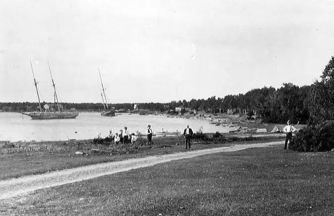 Black-and-white shoreline photograph with sailing vessels, people, and a path beside calm water.