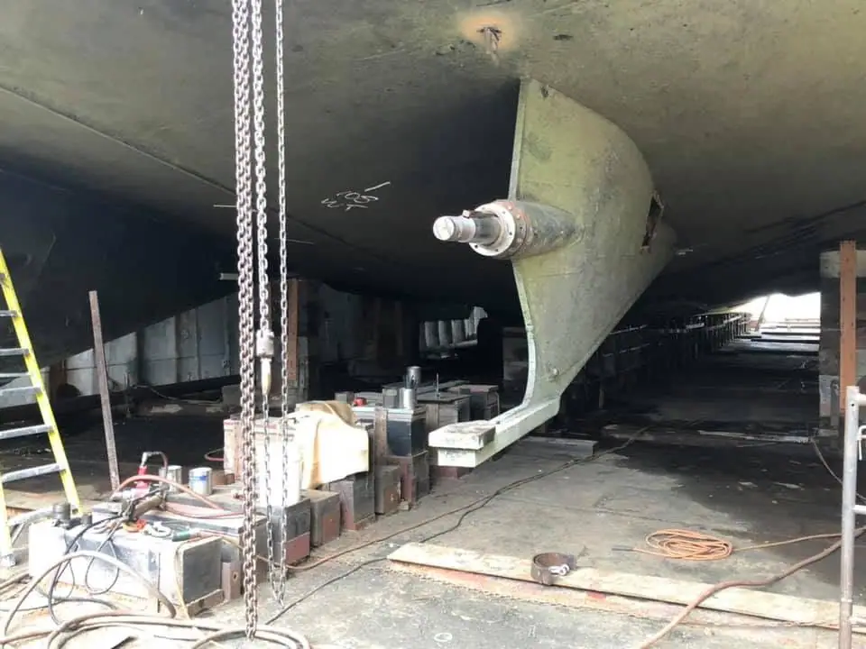 A green-painted vessel hull is raised inside a workshop, with a metal shaft and repair equipment visible.
