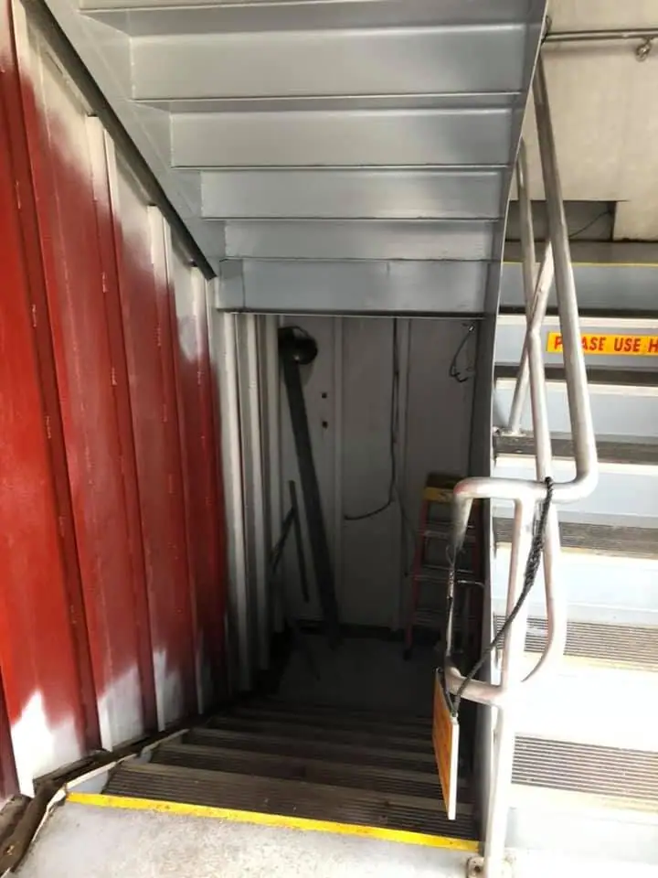 A metal stairway descends into a narrow vessel passage beside a red door and a partially visible safety sign.