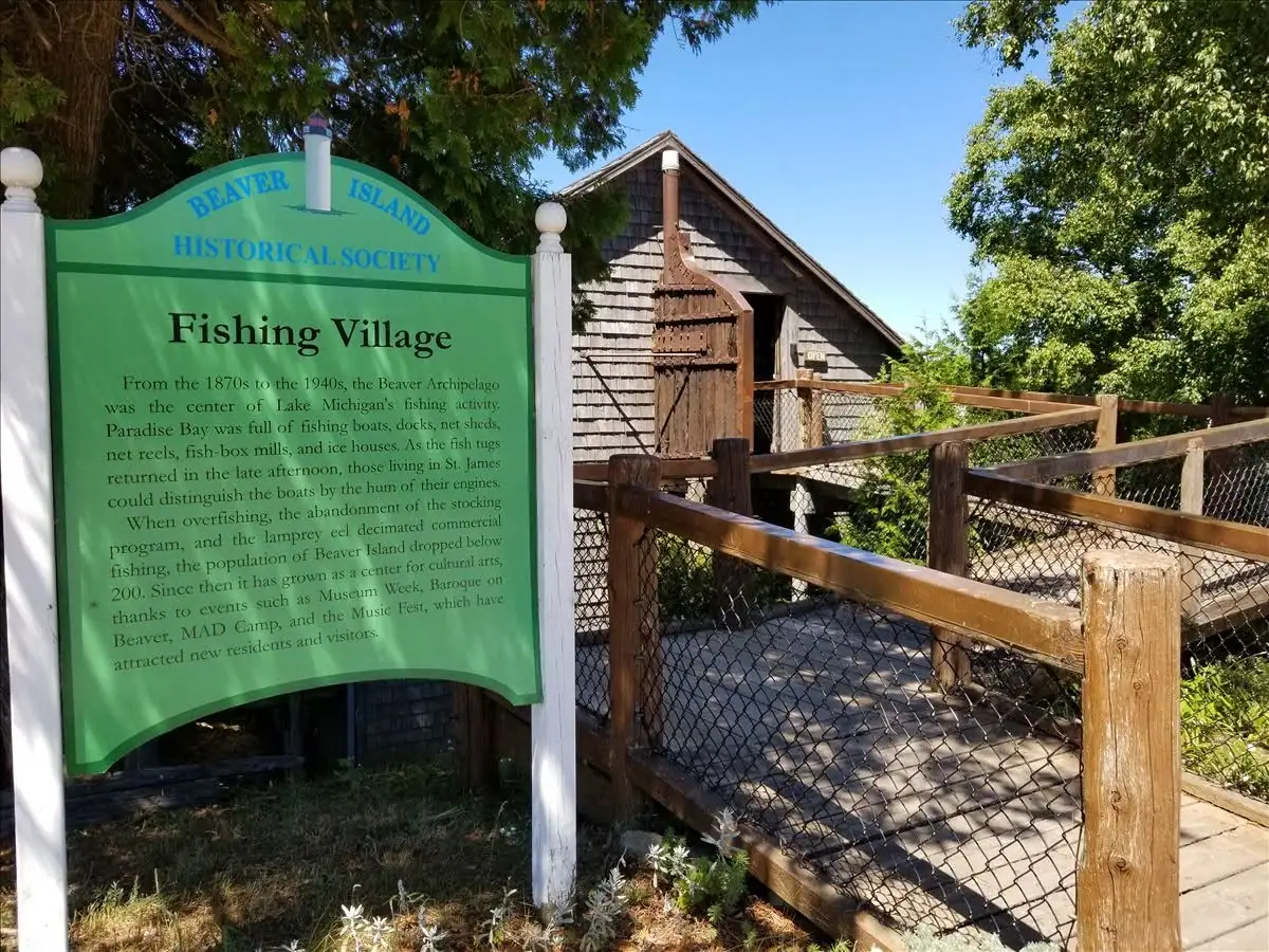 A green historical sign stands beside a weathered shingle-sided building and raised wooden walkway.