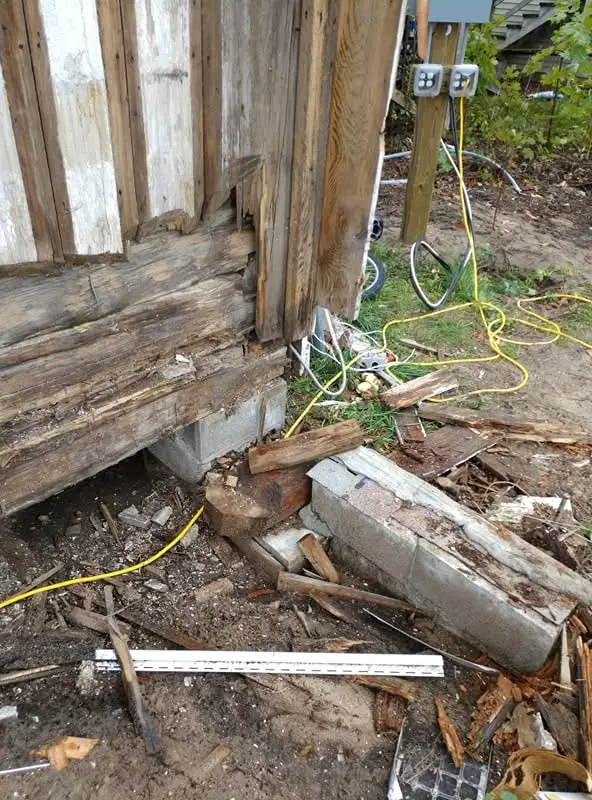 Close view of a rotting wooden structure with electrical outlets, cords, concrete blocks, and scattered debris.