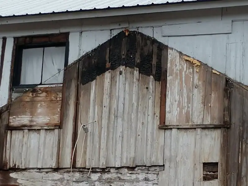 Weathered wooden building facade with a corrugated metal roof, window, boarded openings, and darkened wood.