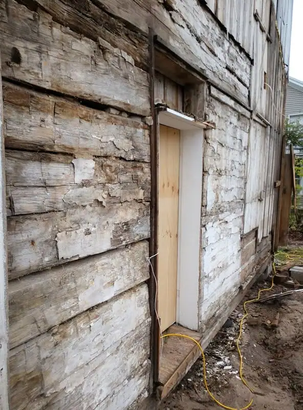 Weathered wooden building exterior with a narrow new door, exposed framing, and a yellow extension cord.