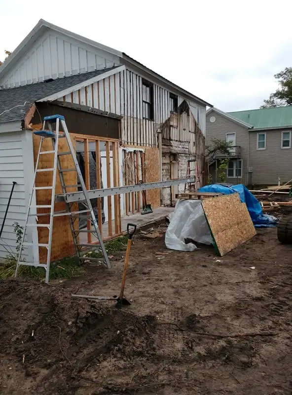 A partially renovated house with exposed framing, a ladder, construction materials, and a shovel in the dirt.