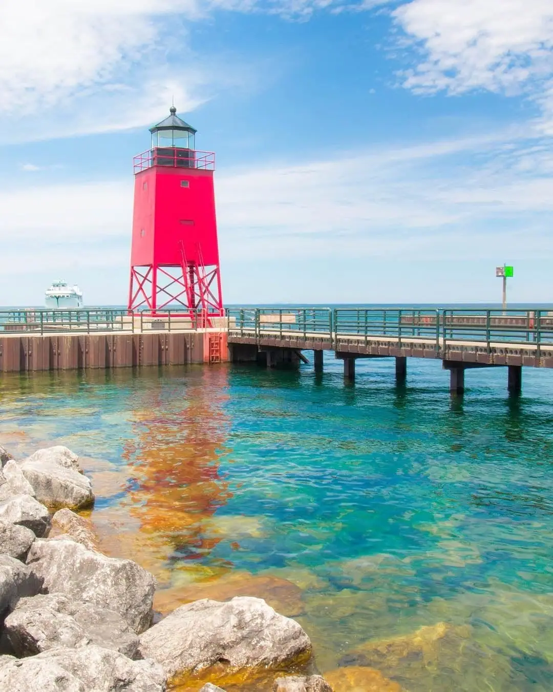 A red lighthouse stands beside a railed pier over clear blue-green water, with rocks in the foreground.