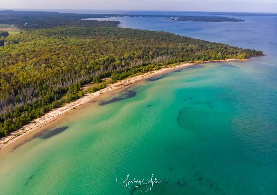 Aerial photograph of a forested shoreline curving around a sandy beach and clear turquoise water.