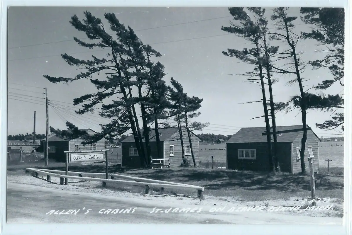Black-and-white view of small wooden cabins beside a road, with pine trees, utility poles, and water behind them.