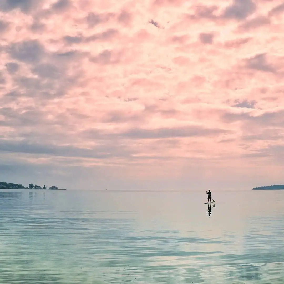 A lone paddleboarder crosses calm water beneath a pastel pink and lavender sky.