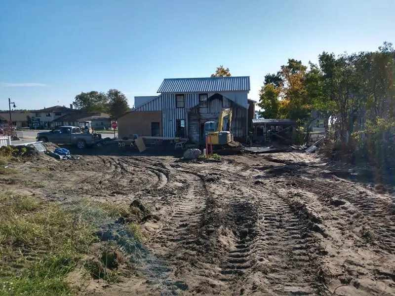 A muddy construction site with a corrugated-metal building, excavator, pickup truck, and deep tire tracks.