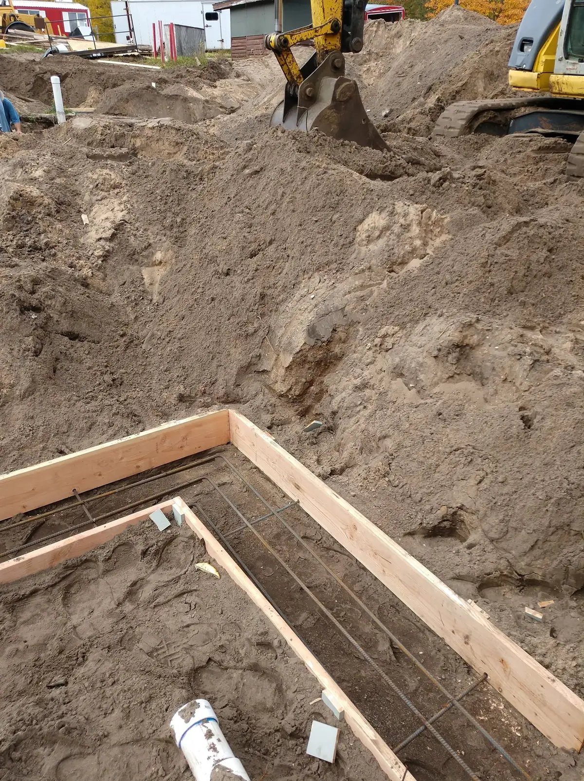 Excavator beside a dirt excavation with wooden foundation forms, steel rebar, and a white PVC pipe.
