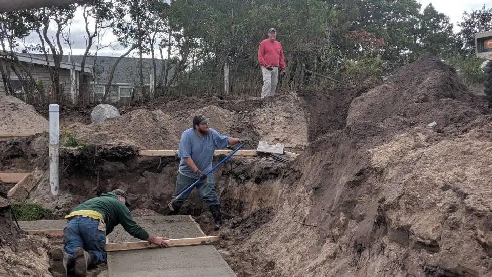 Three workers excavate a trench and finish a rectangular concrete slab beside a house.