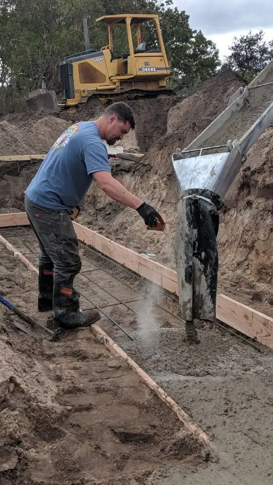 Worker pours concrete into a rebar-reinforced wooden form beside a tracked bulldozer.