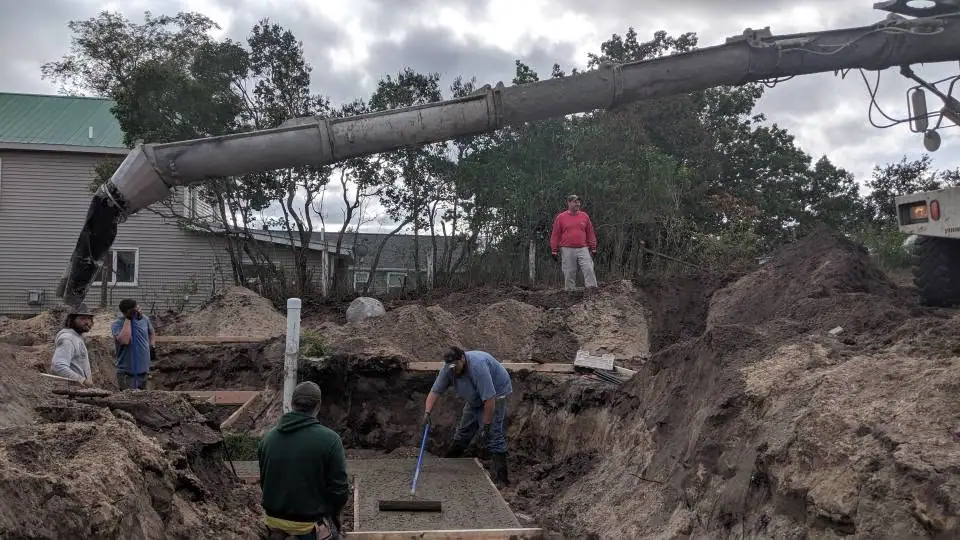 Workers pour and level concrete in an excavated foundation trench beside a house.