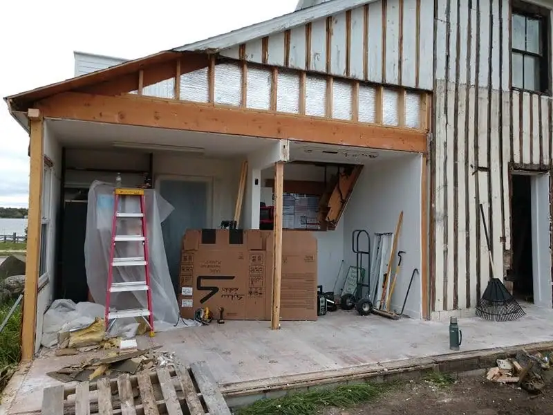 A partially renovated building has exposed framing, a red ladder, boxes, and tools on its covered porch.