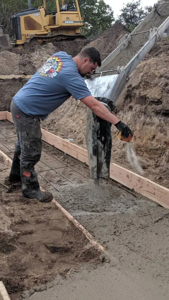 A worker directs wet concrete into a wooden form beside a tracked excavator at an active construction site.