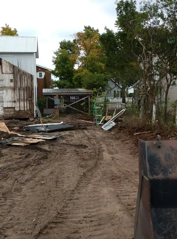 Muddy construction site with a corrugated shed, green ladder, scattered debris, and heavy equipment.