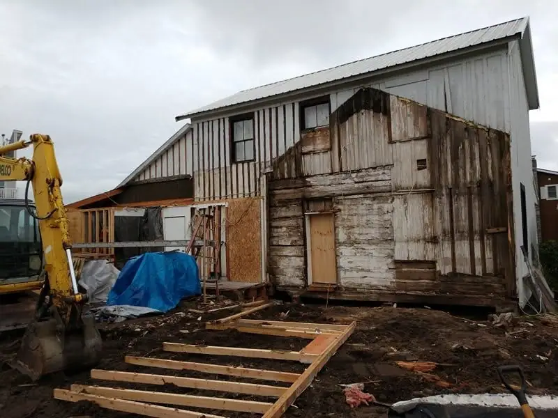 A yellow excavator stands beside a weathered, partially dismantled wooden building with construction debris in front.
