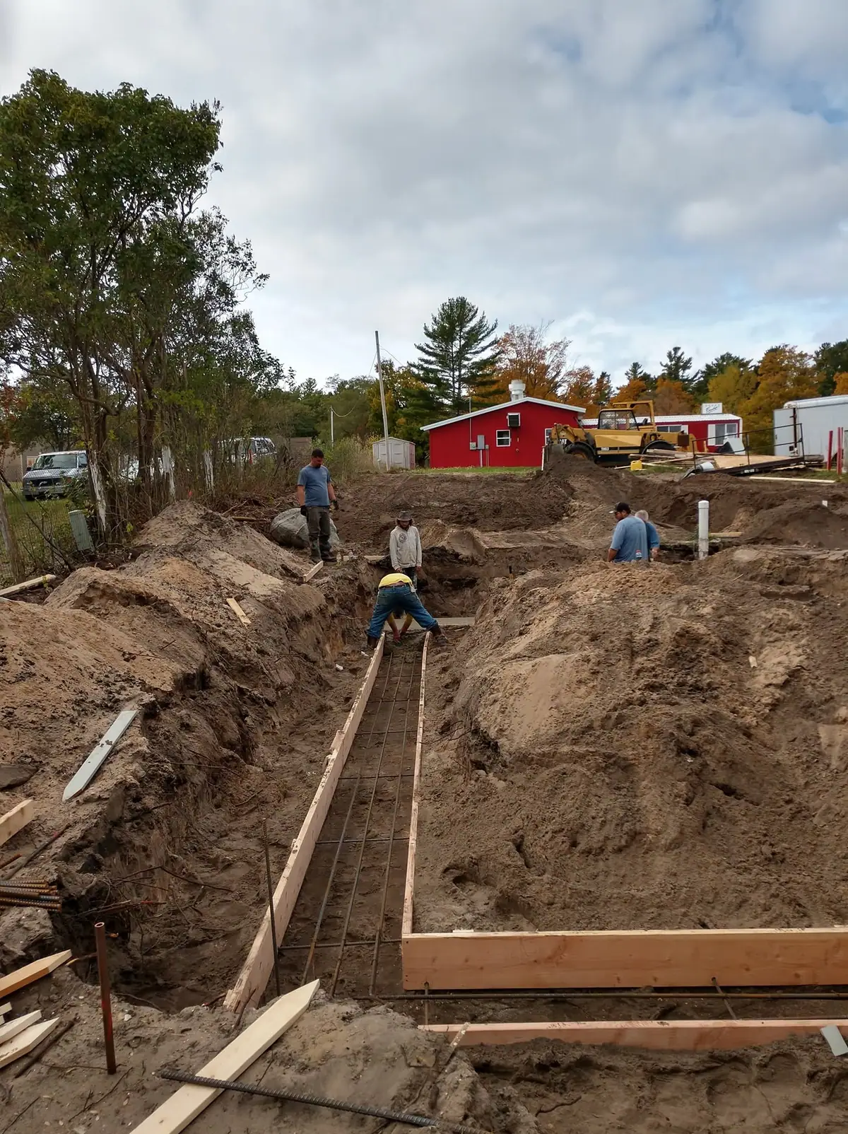 Workers prepare a reinforced concrete foundation in a deep excavation beside a red building and earthmoving equipment.