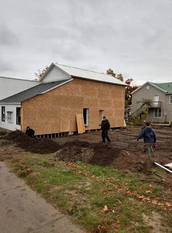 Workers stand beside a partially sheathed building on a muddy lot.