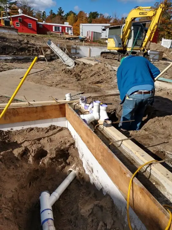 A worker installs pipes in a concrete foundation while an excavator stands behind the construction site.