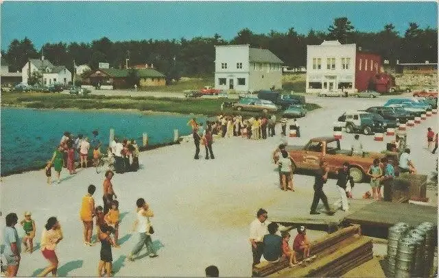 A crowd gathers on a waterfront dock beside parked cars, a red pickup, and buildings along the shore.