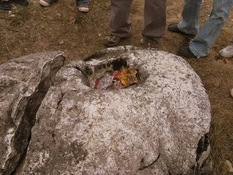 A weathered stone with a central depression holding colorful fabric, surrounded by people standing on dry grass.