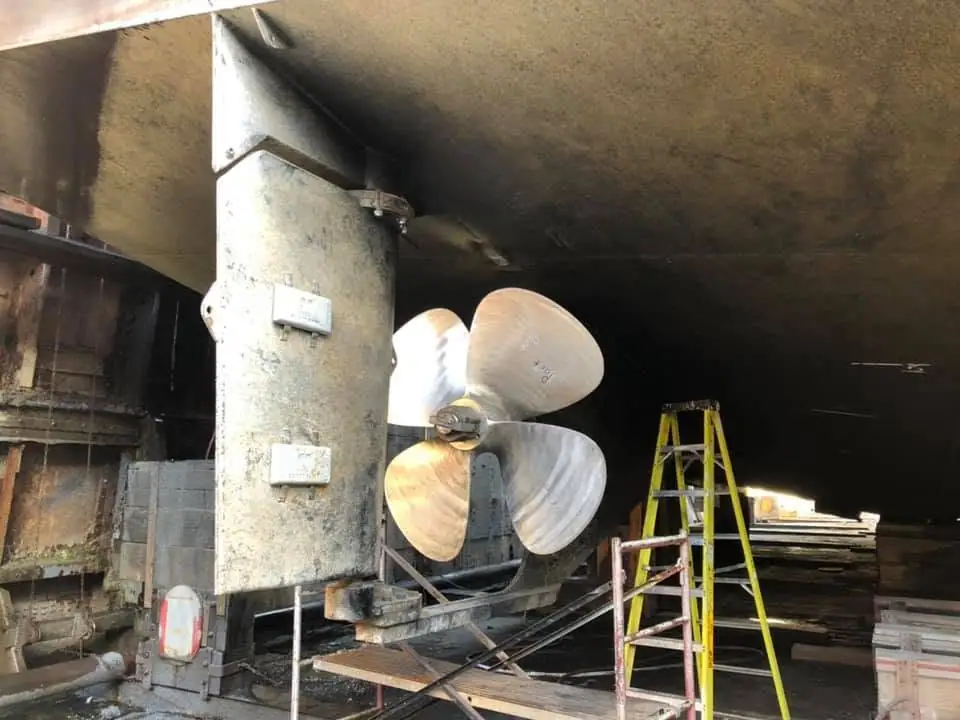 A large marine propeller and shaft stand inside a dim industrial workspace beside ladders and stacked materials.