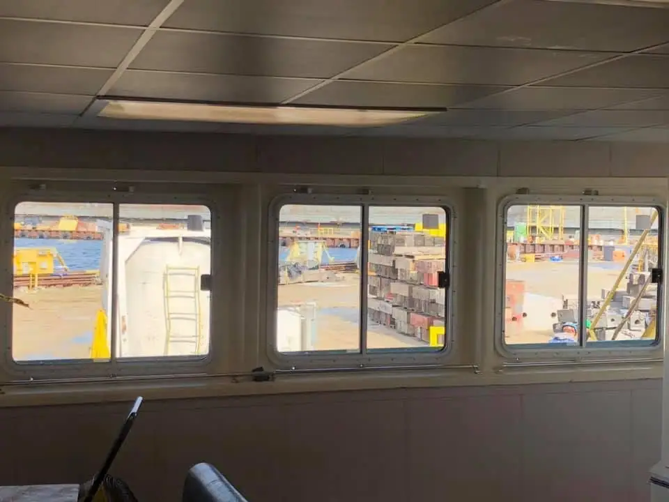 View through a ship's bridge windows toward an industrial waterfront yard with stacked materials and cranes.