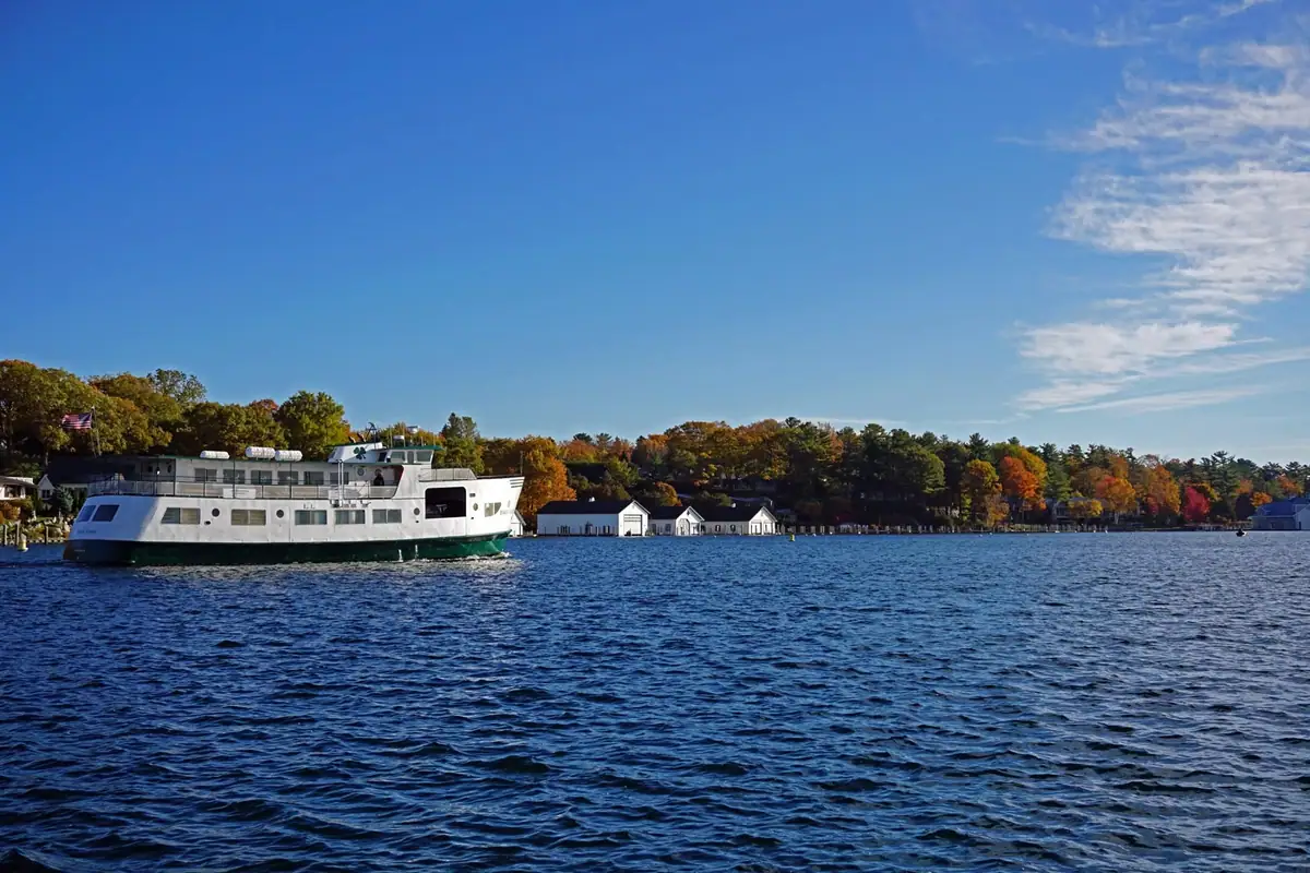 A white ferry crosses dark blue water beside a wooded shoreline with autumn foliage and small buildings.