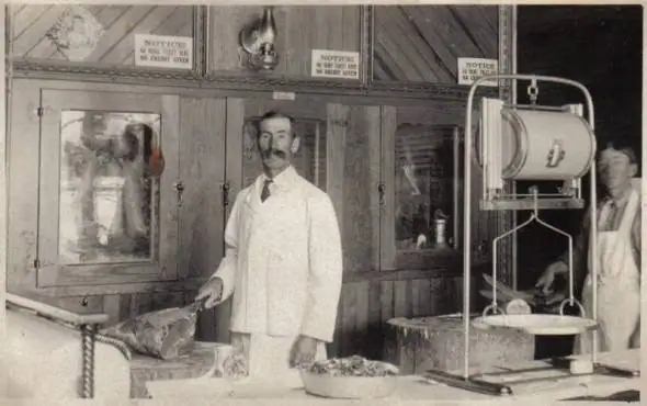 Two men work behind a meat counter, with a large scale and displayed cuts of meat.