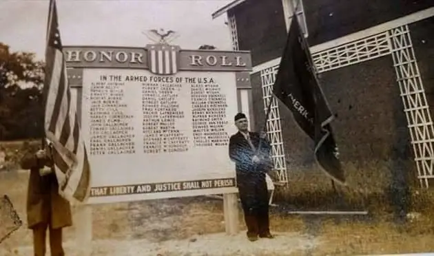 A person stands beside an honor roll monument with American and black flags outside a building.