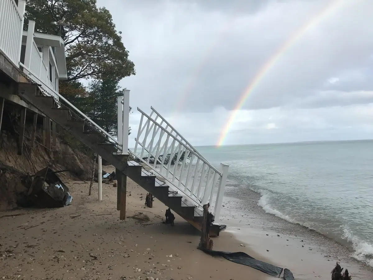 A damaged white staircase descends from a house toward a beach beneath a rainbow and cloudy sky.