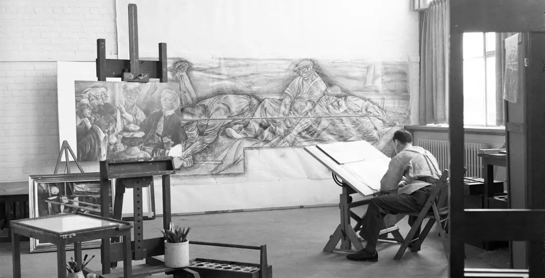 Black-and-white photograph of a person working at a drafting table in a studio with a large mural sketch.