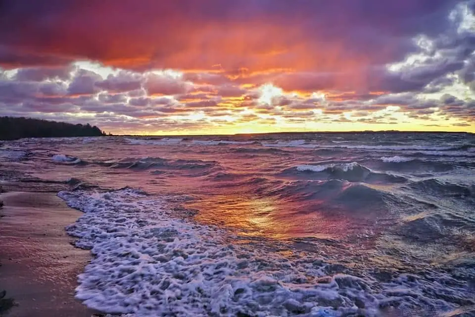 Waves wash a sandy shoreline beneath a vivid, cloud-filled sunset.