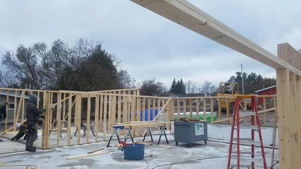 Workers build a timber-framed structure beside an excavator and ladder.