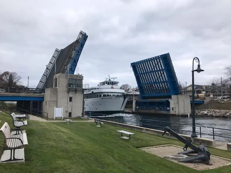 A white ferry passes between the raised spans of a blue drawbridge beside a grassy waterfront.